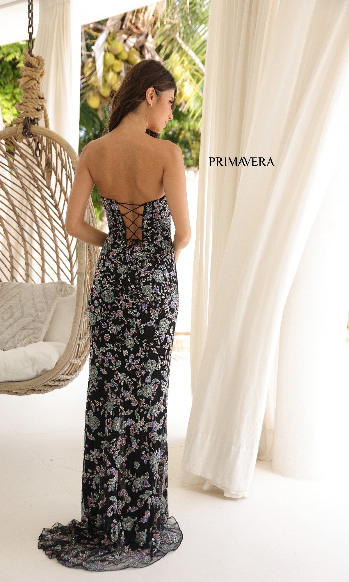 A woman wears the Primavera 4338 Long Sequin-Print Prom Dress, standing indoors near white curtains and a hanging chair.